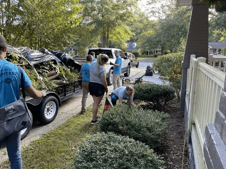 In the picture, a group of volunteers wearing blue shirts is helping to trim bushes at a house with grey painted brick and a white railing. There is also a trailer in the image that is full of branches and limbs.