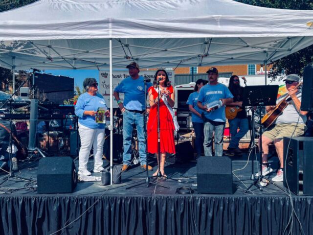 Miss Camden and the United Way are on a stage with a man playing the guitar. The stage is black and there's a white tent.