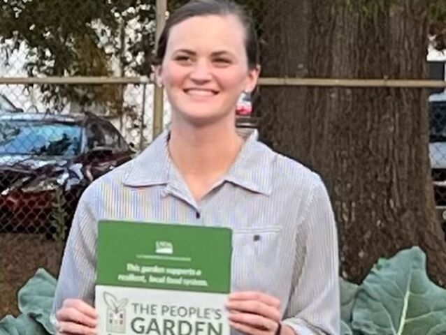 Hanna is holding an award for her work on "The People Garden." She is wearing a shirt with horizontal lines on it. She has her hair tied back in a ponytail.