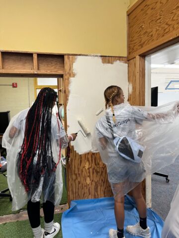 Two individuals, both women with long, thin strands of hair known as braids, are using brushes to apply paint to the surface of a building, which is colored white.