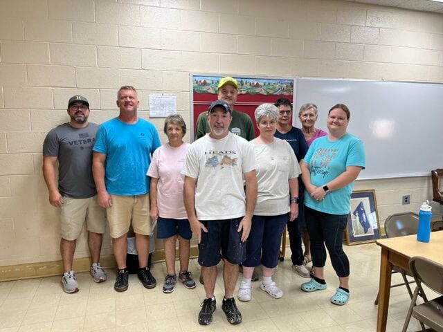 Group photo of elderly individuals gathered in a school setting. The backdrop is a tan cement block wall, and there are tables and chairs positioned in one of the corners.