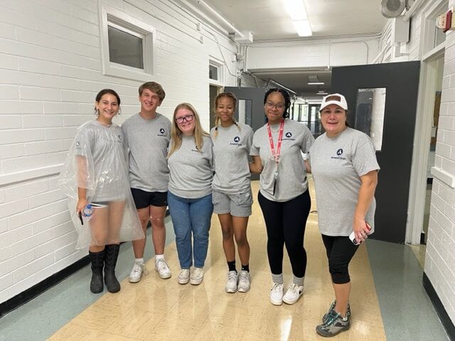 The photo shows a group of volunteers who are helping the community. They are all wearing grey shirts. Some of them are wearing black shorts, some are wearing leggings, and some are wearing jeans.