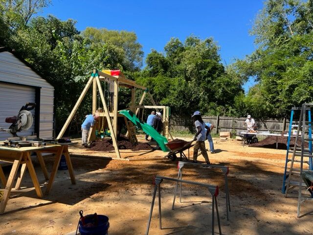 In the picture, there is a volunteer who is helping to put together a wooden playset. Additionally, there is a person pushing a wheelbarrow filled with mulch.