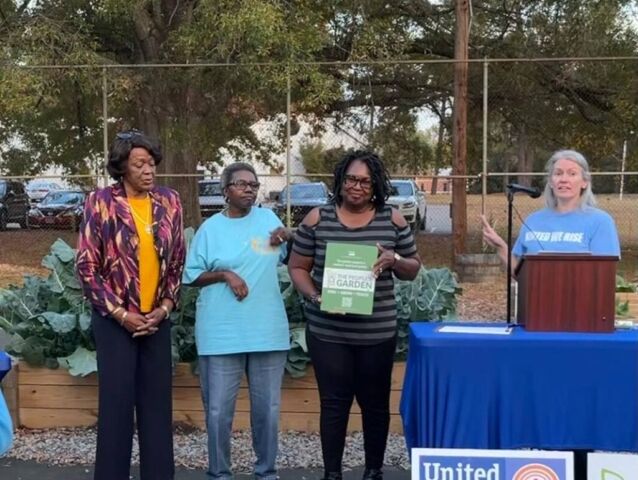 Laura Gunter, a woman, is presenting an award to three individuals of African descent for their contributions to the community garden. In the background, there are garden boxes, adding a peaceful and natural ambiance to the scene.