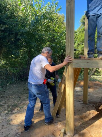 Imagine a playground with a set of steps. Two people are using a drill to attach wooden planks together to make a step for the playset. Meanwhile, someone else is at the top of the steps, doing something else.