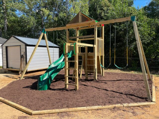 The picture shows a wooden playset with a soft, spongy ground. In the background, there are tin sheds.